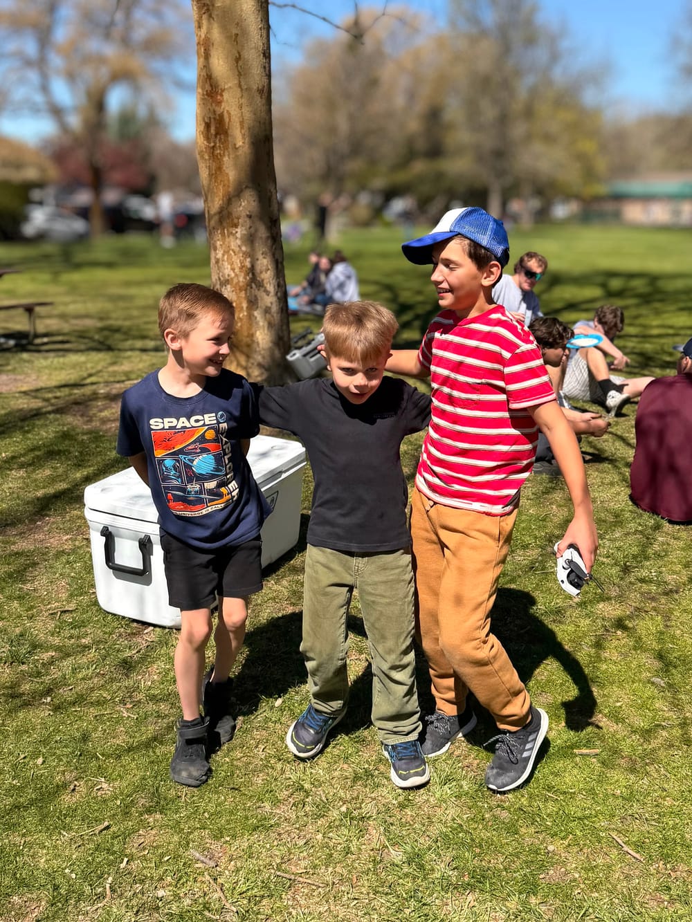 Three young boys with arms around each other at a Morning Star Christian Church community outreach event in a Boise park, summer afternoon